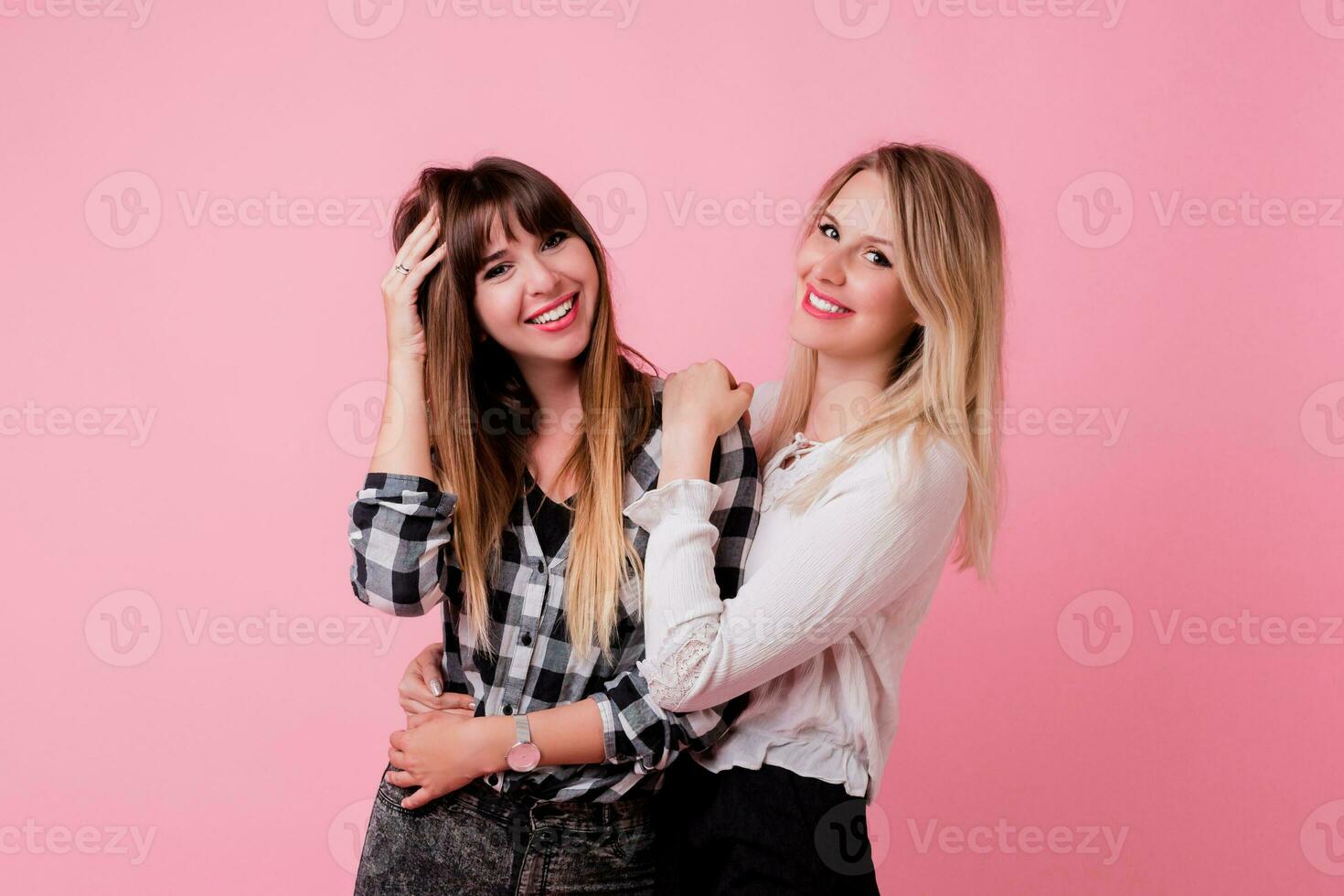 Two smiling women hugging and standing isolate over pink background . Brunette and blonde girls. Natural make up. photo