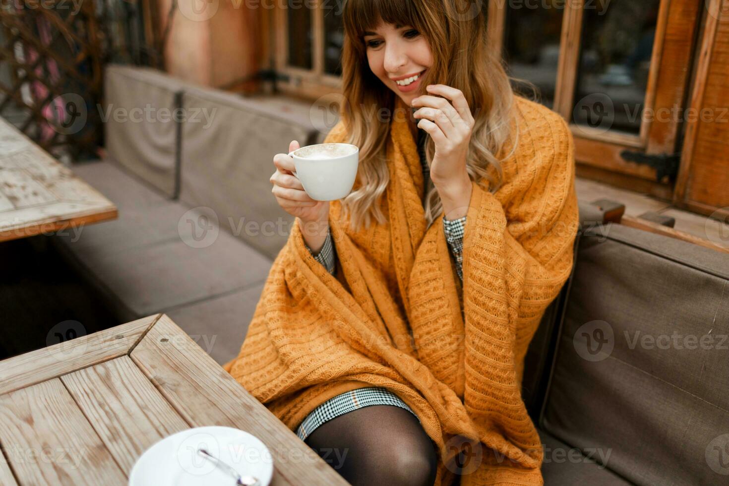 Warm cozy portrait of happy dreamy woman with wavy hairs, covered by  yellow  plaid and holding cup of hot cappuccino.  White female chilling on terrace. photo