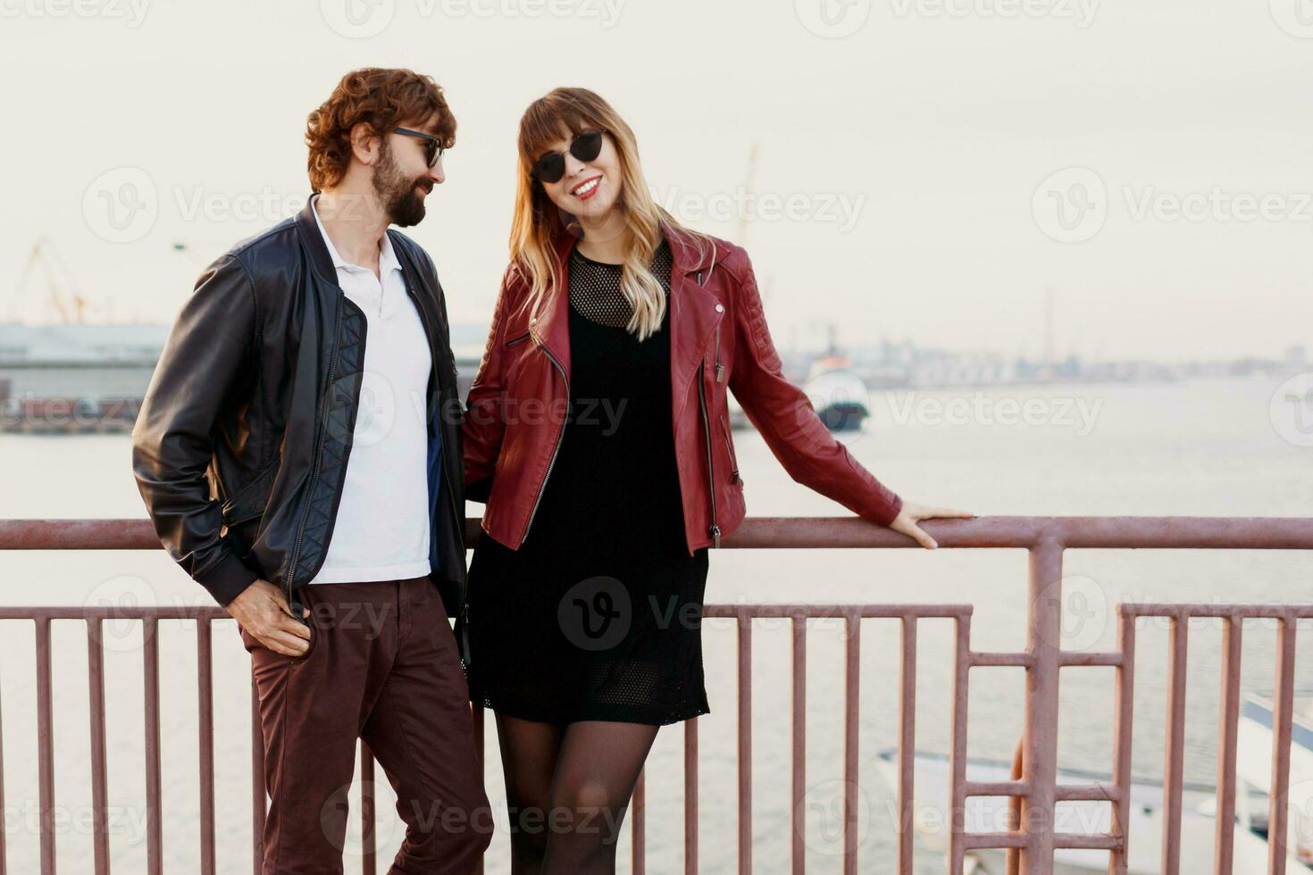 Outdoor fashion image of  stylish couple in casual outfit, leather jacket and sunglasses standing on the bridge.   Handsome man with beard with his girlfriend spending romantic time together. photo