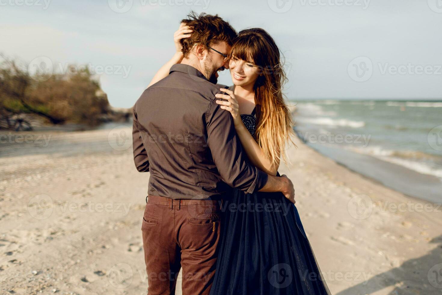 Two happy people in love, elegant woman with her handsome boyfriend hugging and enjoying romantic time together on the beach. Elegant dress. photo