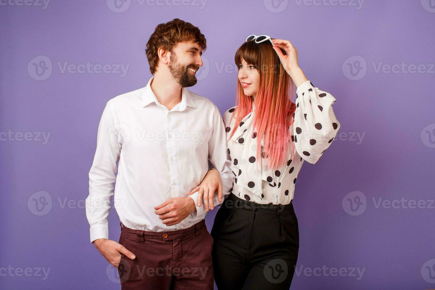 Cute couple hugging and looking on each other.  Handsome  man with beard and his pretty woman with pink hairs in stylish retro outfit posing on purple background in studio. photo