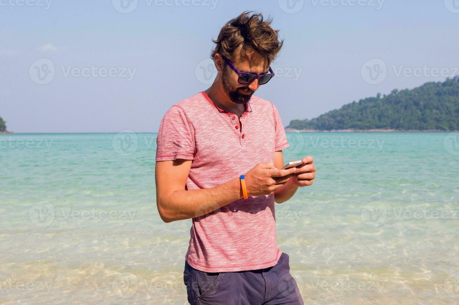 Stylish man with beard  using smartphone on his tropical vacations.   Sunny  beach background.  Traveler hipster. Holiday work. Sending message. photo