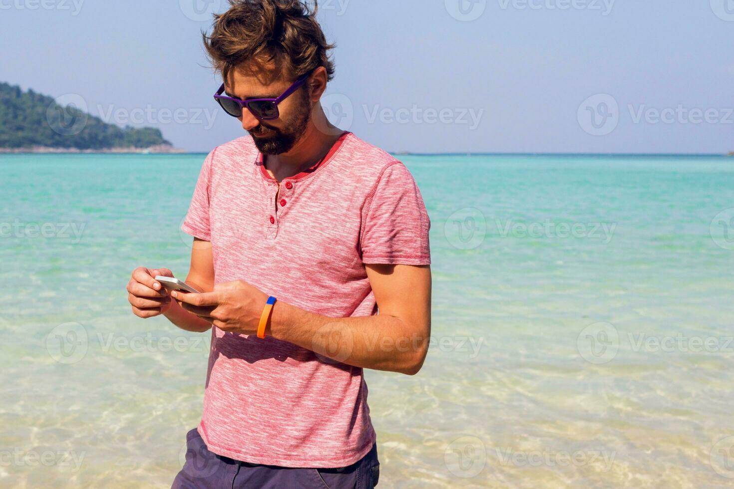 Stylish man with beard  using smartphone on his tropical vacations.   Sunny  beach background.  Traveler hipster. Holiday work. Sending message. photo