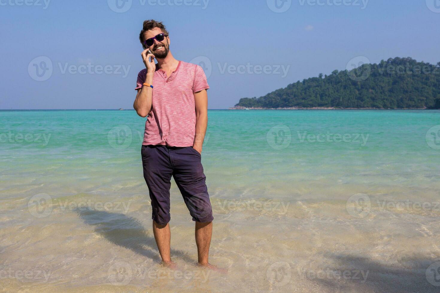 Happy  traveling man with beard  talking by mobile phone and staying  in blue sea water on his tropical  holidays. Holding  phone in hands. Full length. photo