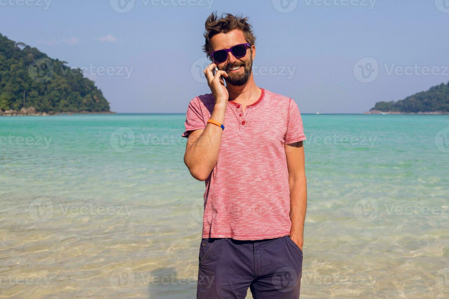 Stylish man with beard  using smartphone on his tropical vacations.   Sunny  beach background.  Traveler hipster. Holiday work. Sending message. photo