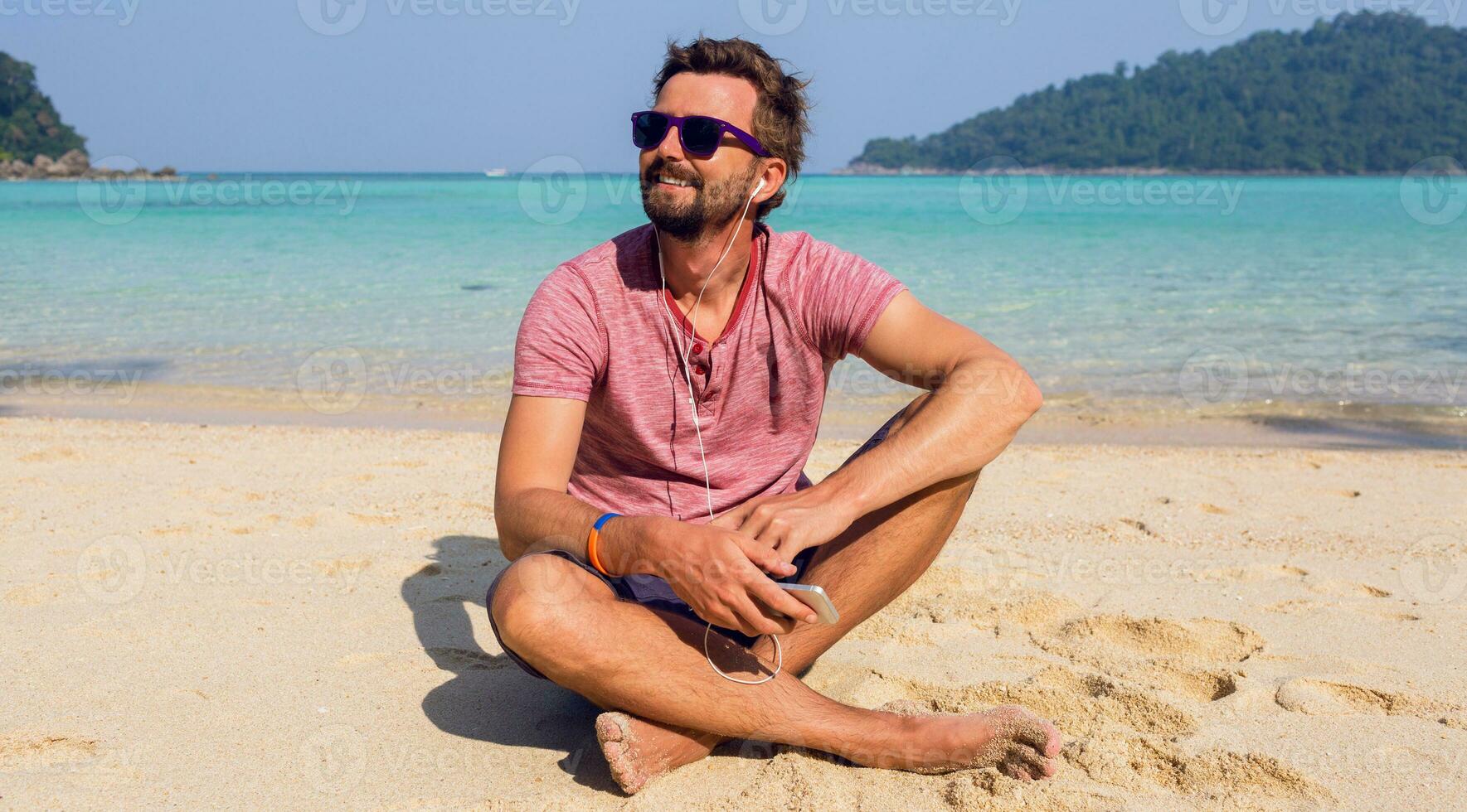 Happy man relaxing on sunny beach, sitting on sand, holding mobile phone. Listening  favorite music. photo