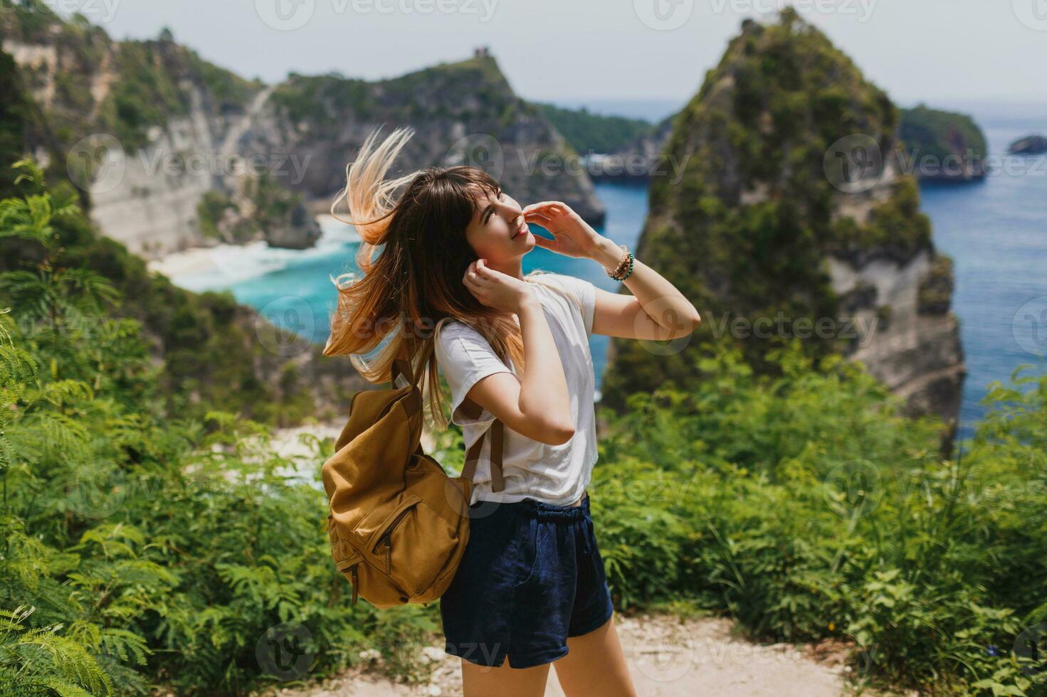 Traveling and adventure concept. Happy pretty woman with back pack  traveling in Indonesia on Nusa Penida island. Amazing cliffs and tropical beach view on background. photo