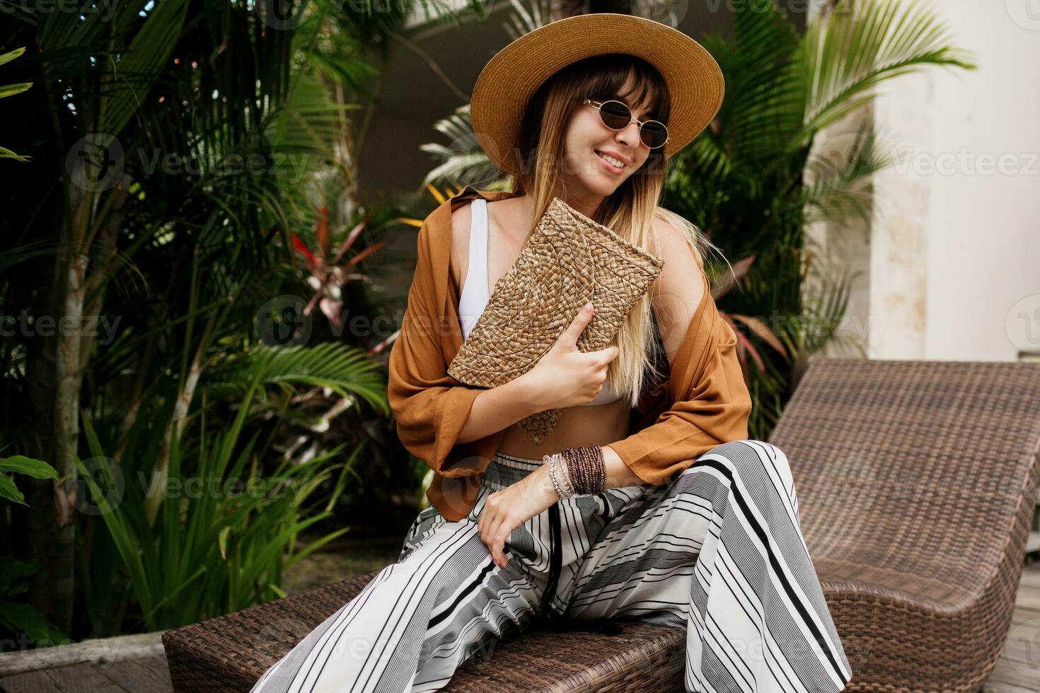 Stylish woman in summer clothes relaxing in hotel and enjoying Trendy sunglasses, straw hat and handbag, bohemian bracelets and accessorizes. photo