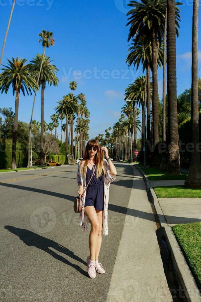 lleno longitud retrato de elegante sonriente mujer caminando en exótico calle cerca el hotel en soleado caliente día . gasto su vacaciones en los angeles foto