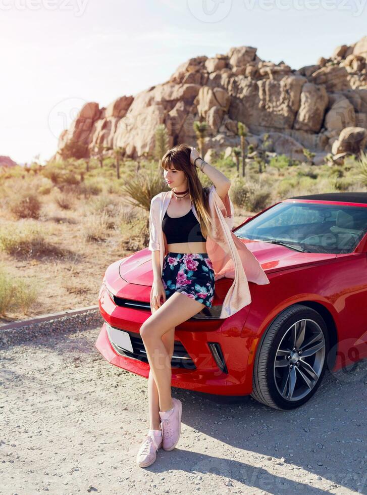 Fashionable image of pretty seductive girl sitting on  red modern sport convertible car. Joshua tree national park background. photo