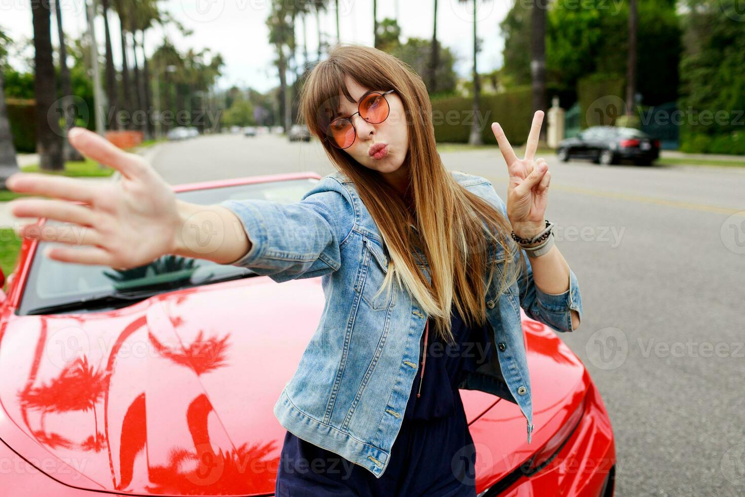 Cute woman in pink glasses making self portrait near amazing red convertible  sport car in California. photo