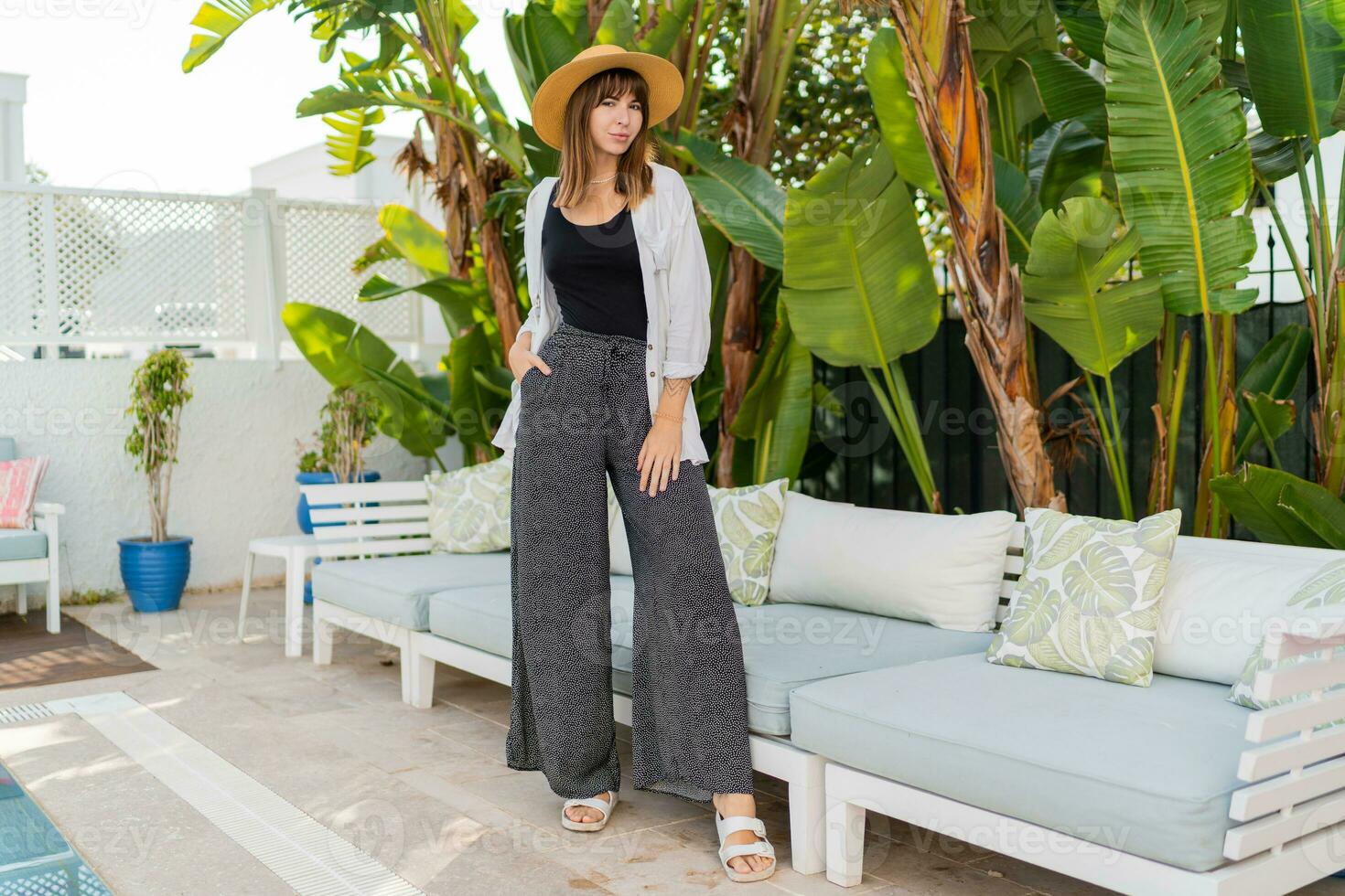 Elegant woman in straw hat posing  in cozy tropical resort near pool. Tropical leaves on background. Stylish white furniture. Full lenght. photo