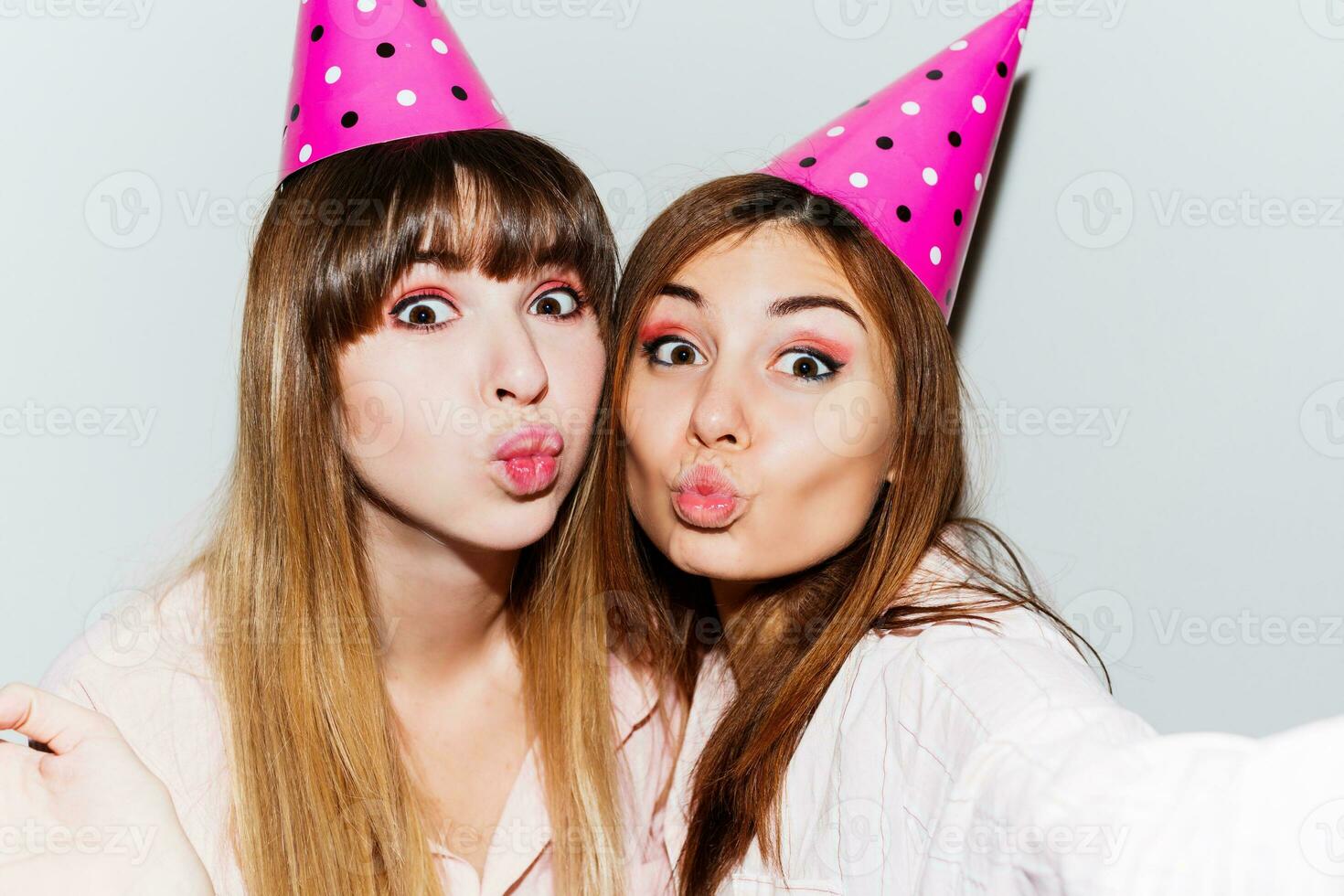 Self portrait of two  women in pink  paper birthday hats  on white background.  Friends wearing pink pajamas and send kiss to camera. Playful mood. photo