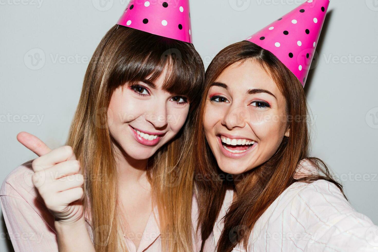 Self portrait of two  women in pink  paper birthday hats  on white background.  Friends wearing pink pajamas and send kiss to camera. Playful mood. photo