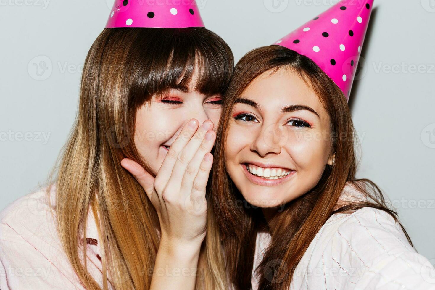 Self portrait of two  women in pink  paper birthday hats  on white background.  Friends wearing pink pajamas and send kiss to camera. Playful mood. photo