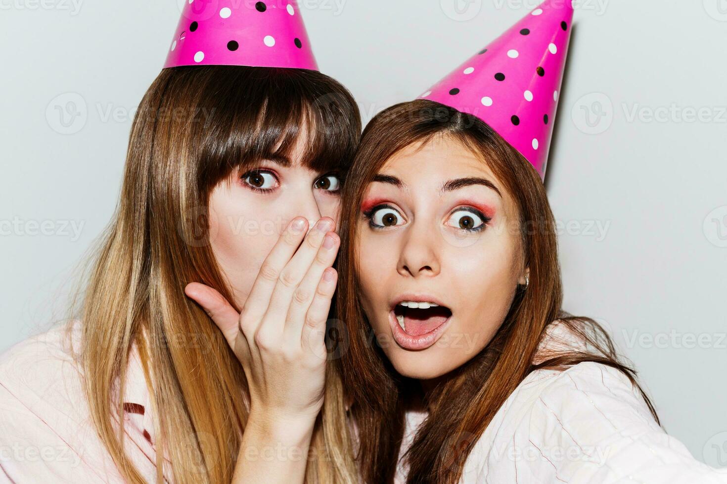 Self portrait of two  women in pink  paper birthday hats  on white background.  Friends wearing pink pajamas and send kiss to camera. Playful mood. photo