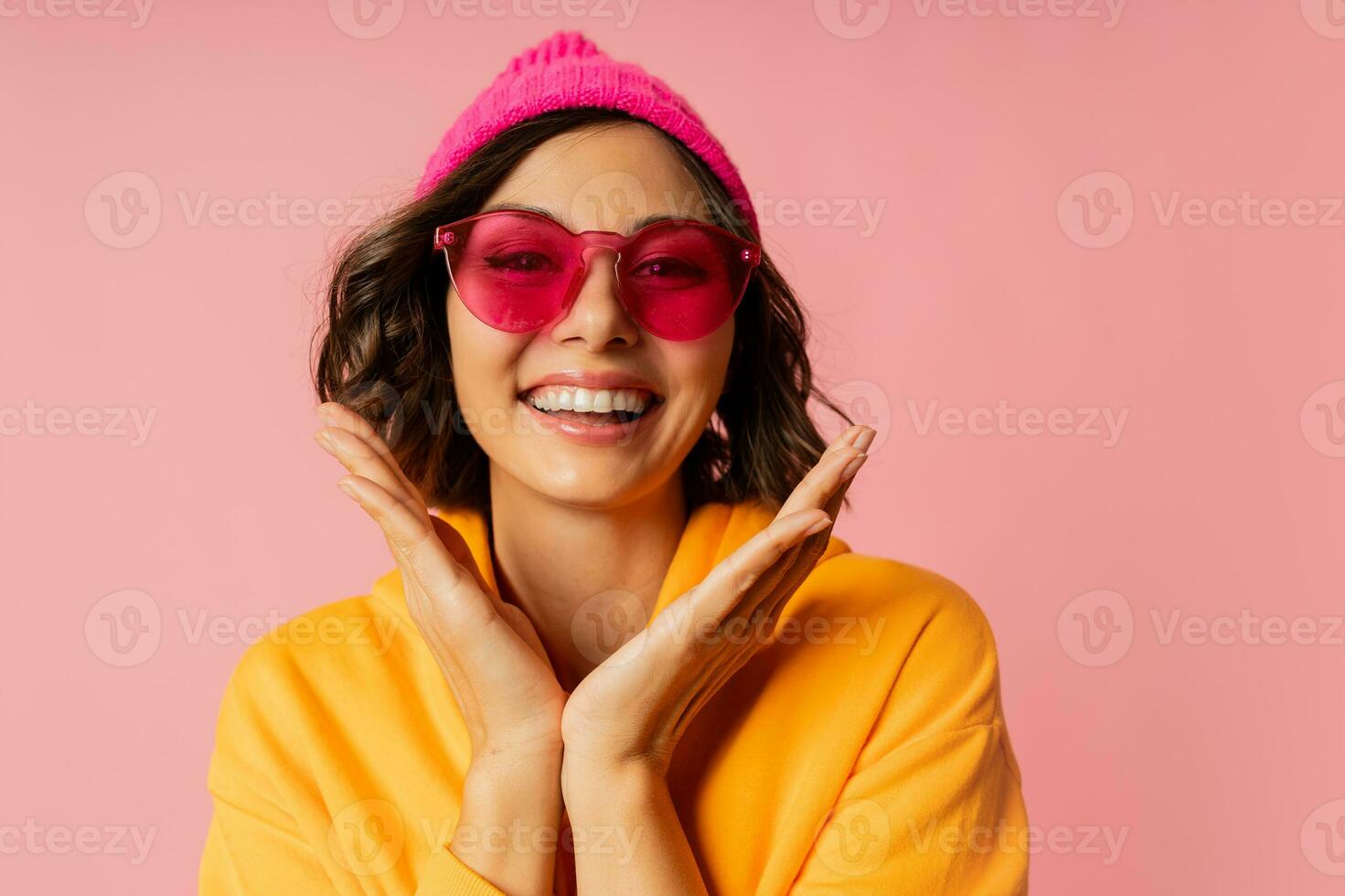 Woman in pink hat and  orange hoodie  with emotional face posing on pink bacground.  Stylish sunglasses. photo