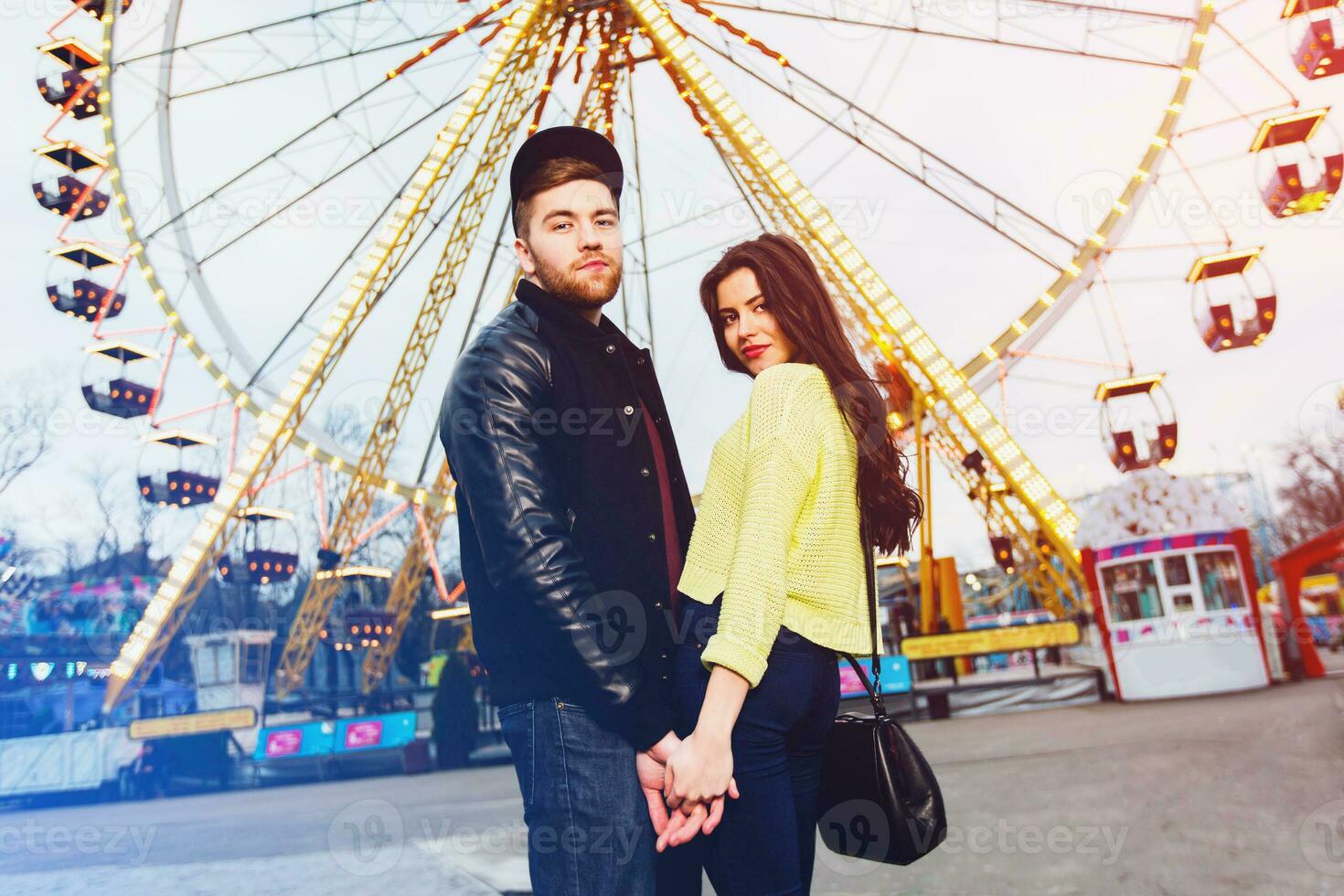 Stylish hipster  couple in love holds hands and enjoying together being in an amusement park  at night in  Odessa . Handsome man and beautiful brunette wearing trendy fall outfit. photo