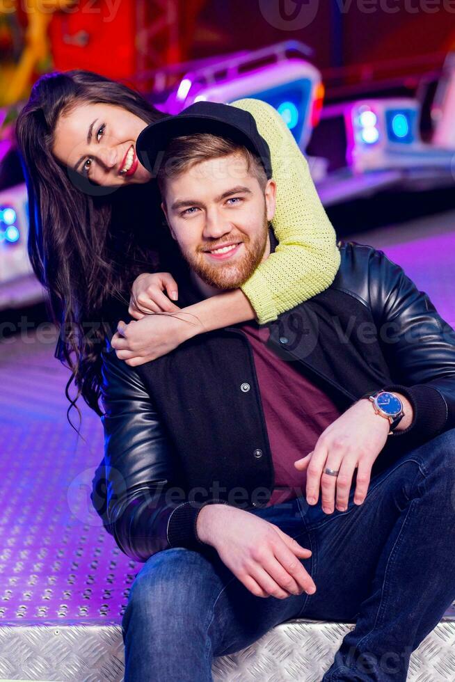 Stylish hipster  couple in love holds hands and enjoying together being in an amusement park  at night in  Odessa . Handsome man and beautiful brunette wearing trendy fall outfit. photo