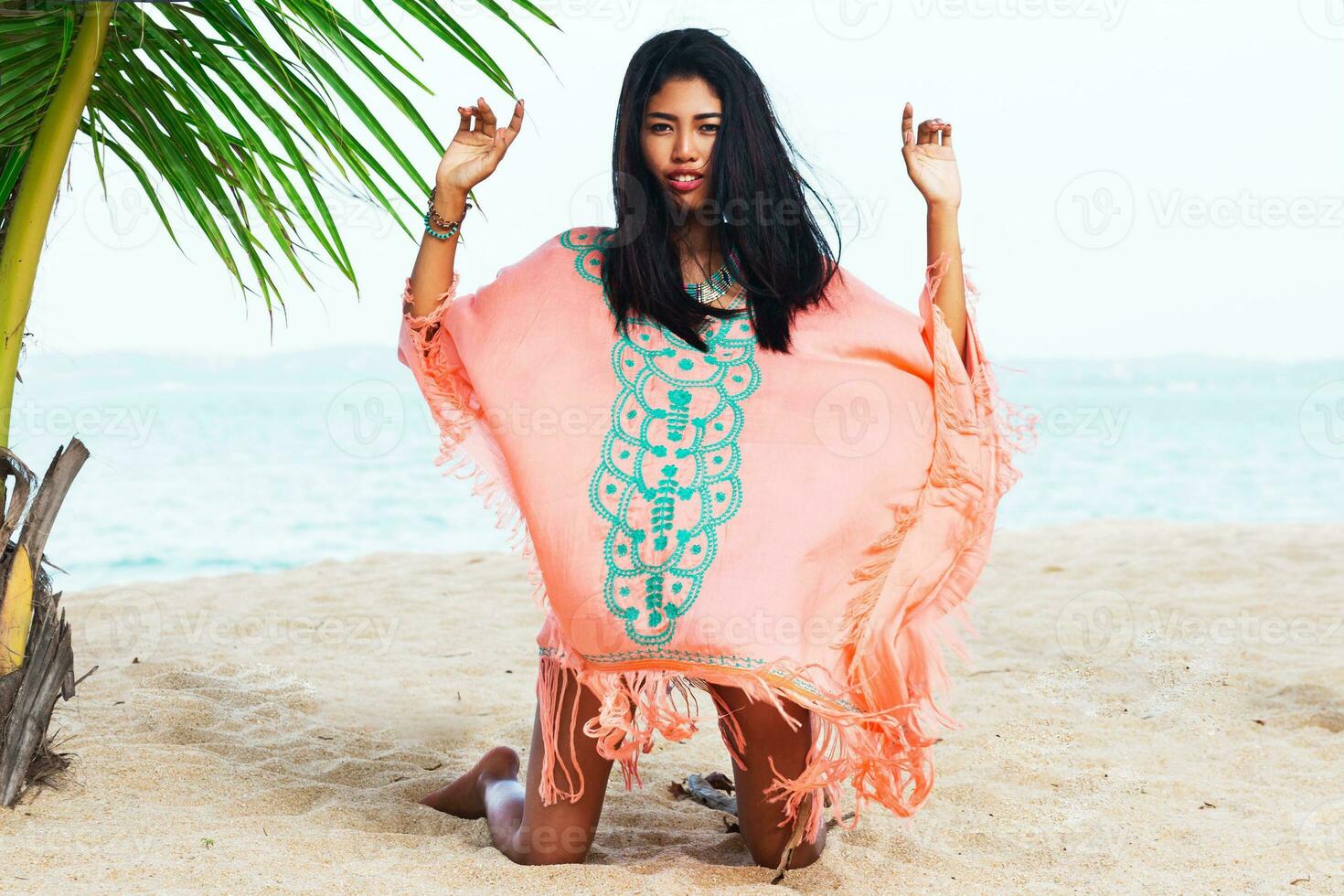 Young  beautiful  asian woman  relaxing on tropical beach, wearing  boho trendy dress, sitting on white sand near  palm tree.   fashion look. photo