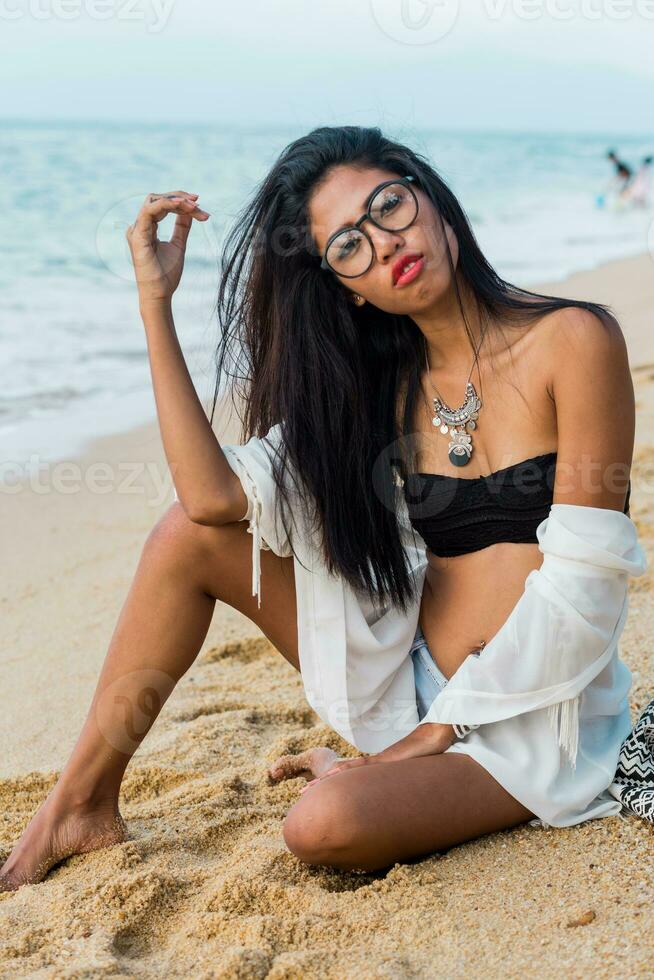 Close up lifestyle portrait of stylish Asian woman with red lips. She is sitting on sunny tropical beach. Boho accessories.Jewelry ,bracelet and necklace. photo