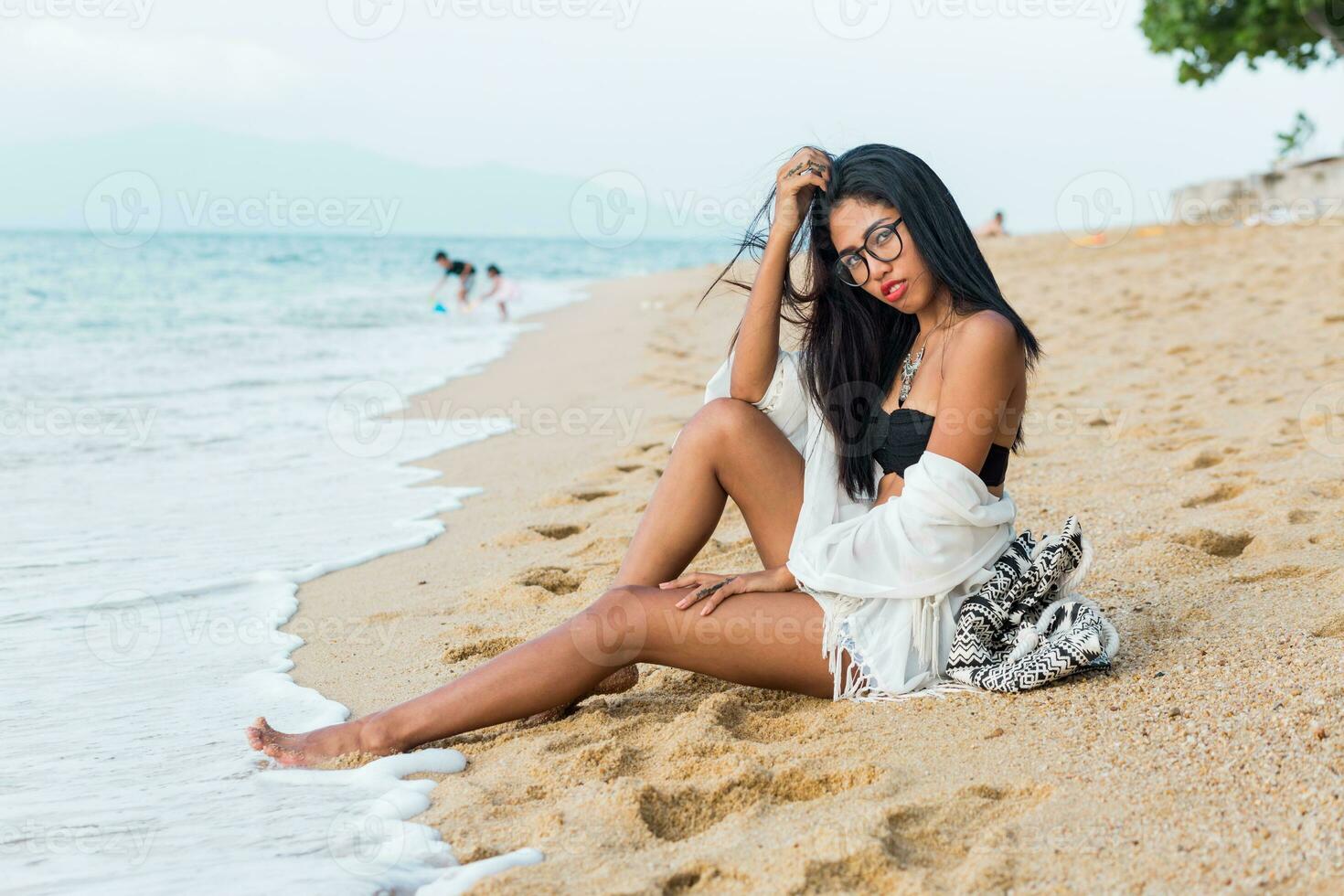 Asian woman with red lips posing on tropical beach in the evening. Wearing boho white cover up, back pack with stylish print. Perfect tan body. Stylish Sumer outfit.Jewelry ,bracelet and necklace. photo