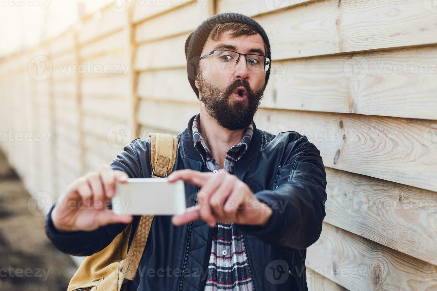 Surprised face. Hipster man making selfie on smartphone, crazy emotions . Posing on wood wall background. Wearing stylish black hat, leather jacket  and backpack. photo