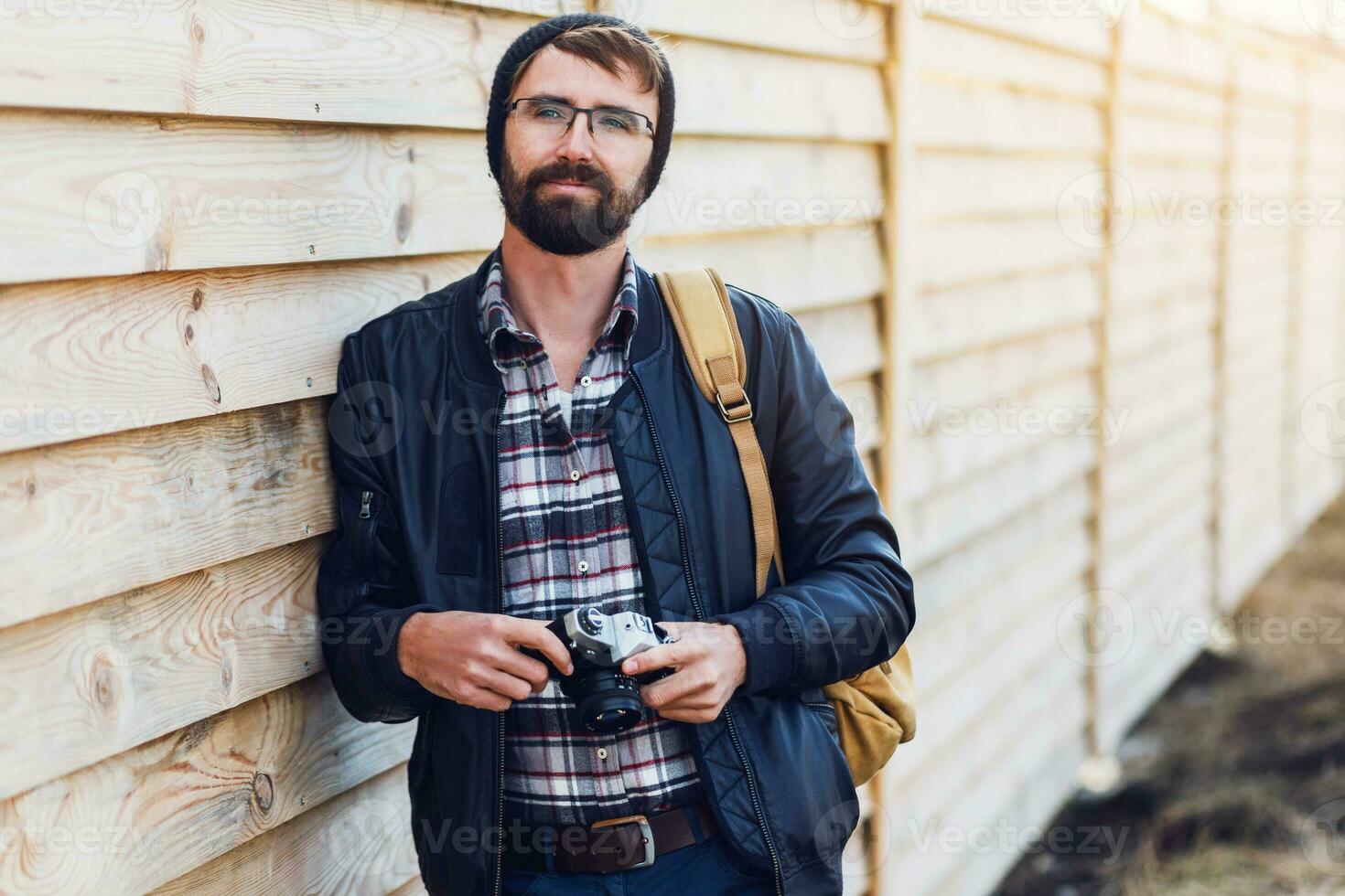 Handsome hipster man with beard, in stylish hat and glasses posing with retro camera in hands  and backpack stands on wooden  background. photo