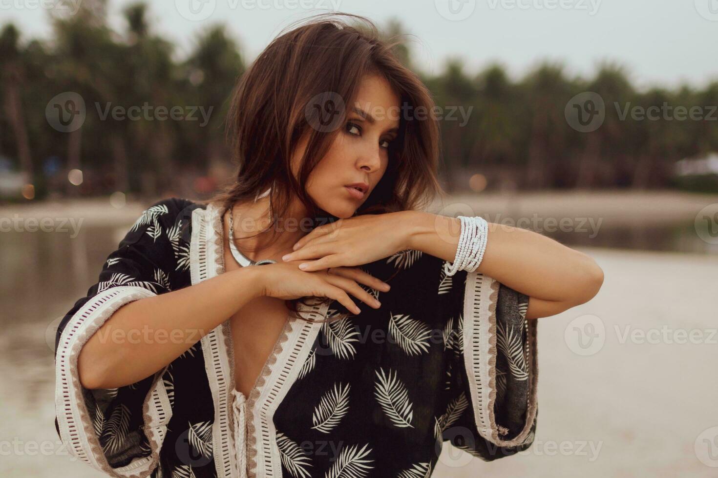 Beautiful Asian woman in tropical bohemian dress posing on the beach. Wearing earrings with feather, necklace and bracelets. photo