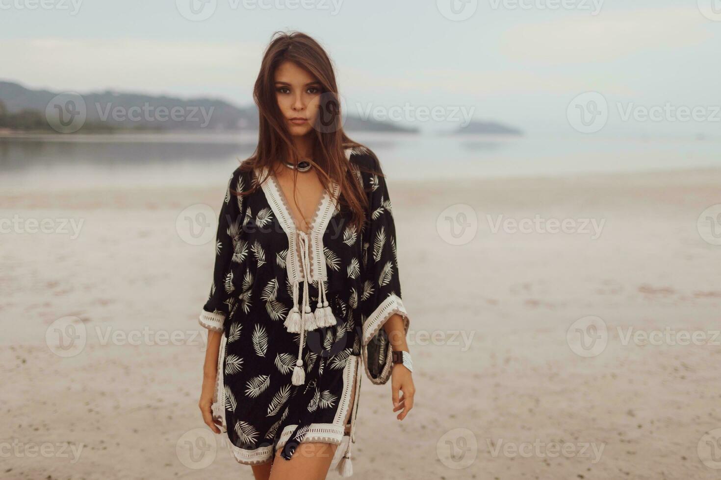 Beautiful Asian woman in tropical bohemian dress posing on the beach. Wearing earrings with feather, necklace and bracelets. photo