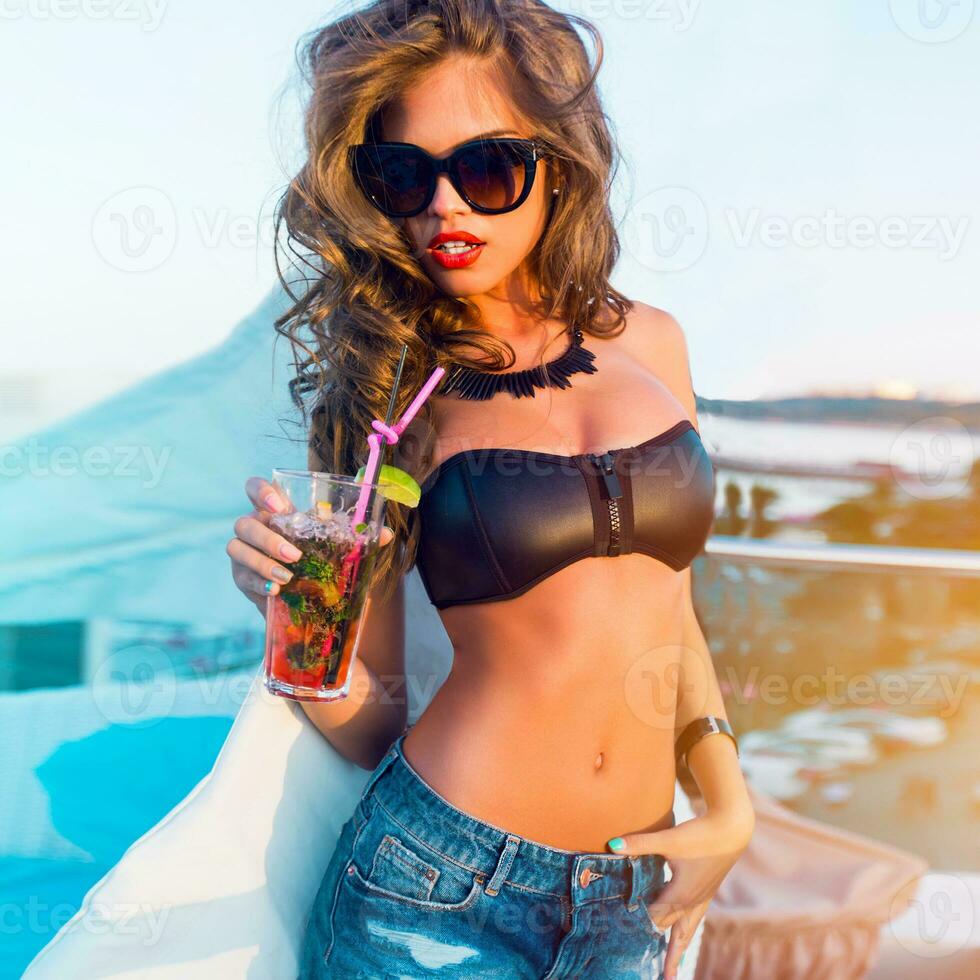 Outdoor summer lifestyle portrait of stylish  sexy   woman with perfect  tanned fit body   wearing trendy sunglasses drinking cocktail and  enjoying  pool party on luxury villa. photo