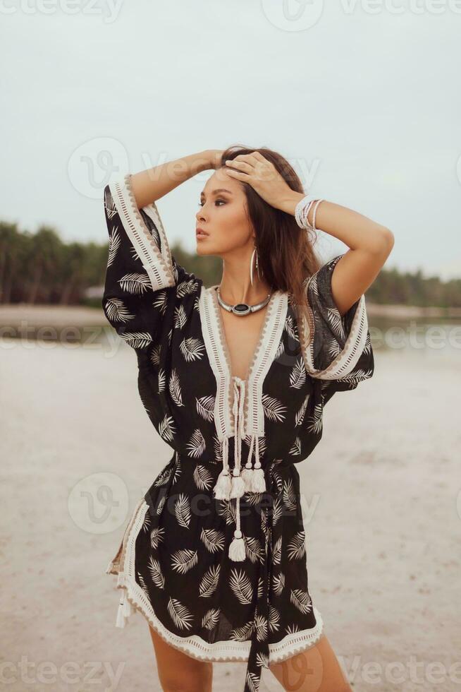 Beautiful Asian woman in tropical bohemian dress posing on the beach. Wearing earrings with feather, necklace and bracelets. photo