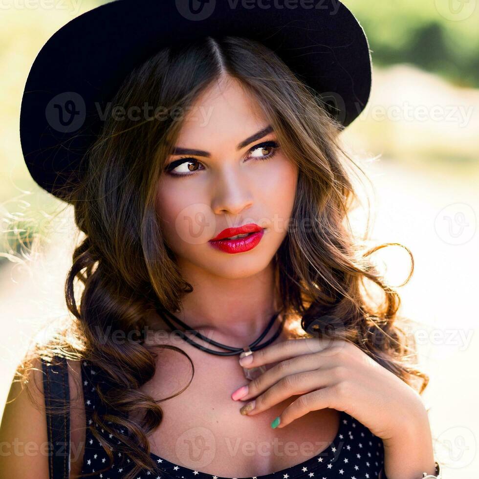 Summer sunshine portrait of pretty young  hipster woman with dark curly hair   in  stylish elegant black hat and bright make up posing in the park.  Street style. photo