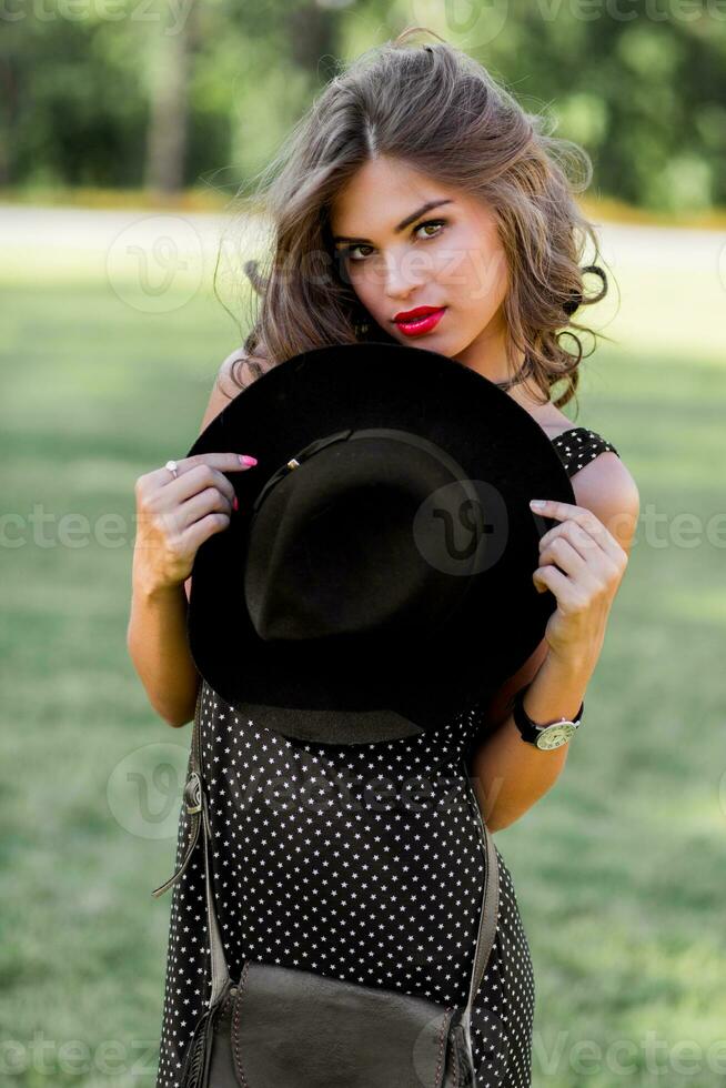 Summer fashion  portrait of r elegant woman with  perfect wavy  hair   in  stylish elegant black hat and bright make up posing in the park.  Street style. photo