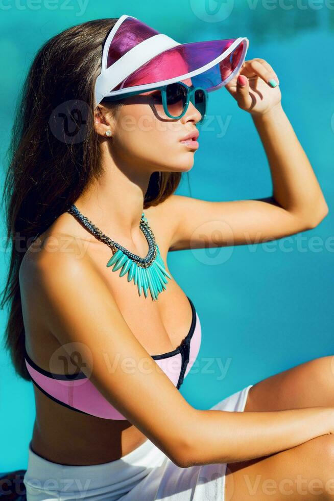 Lifestyle portrait of tanned beautiful woman in  pink  bikini and sunglasses sitting near  swimming pool with  fresh cocktail. Amazing sensual girl with perfect  slim sport body posing outdoor. photo