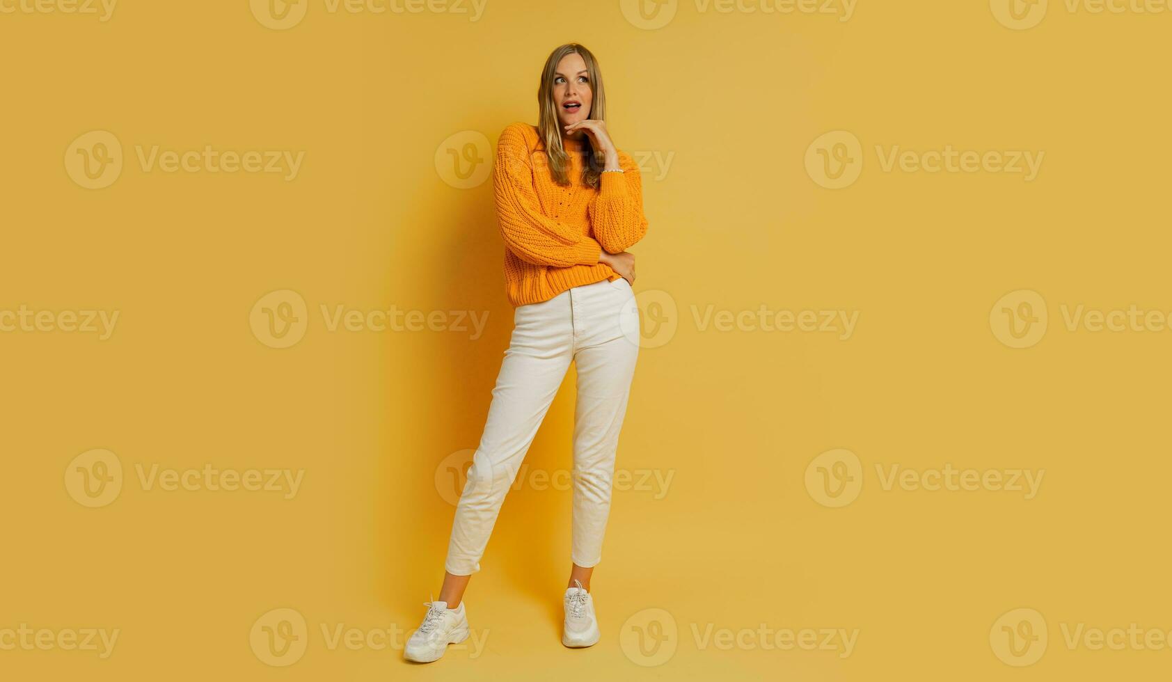 Blond  woman  with suprice face in orange  stylish autumn sweater posing over yellow background in studio.  Full lenght. photo