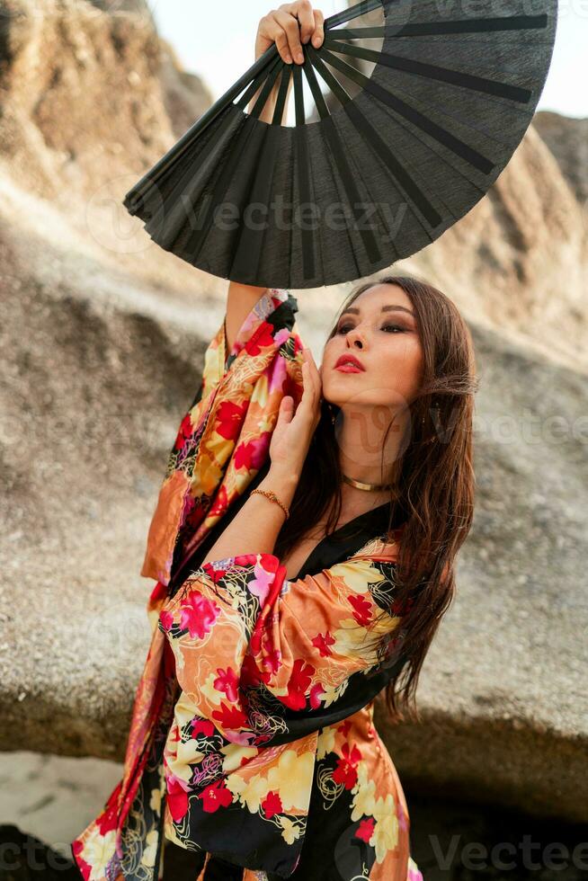 Moda foto de elegante asiático mujer en seda kimono participación ventilador y posando terminado rocas en el playa.