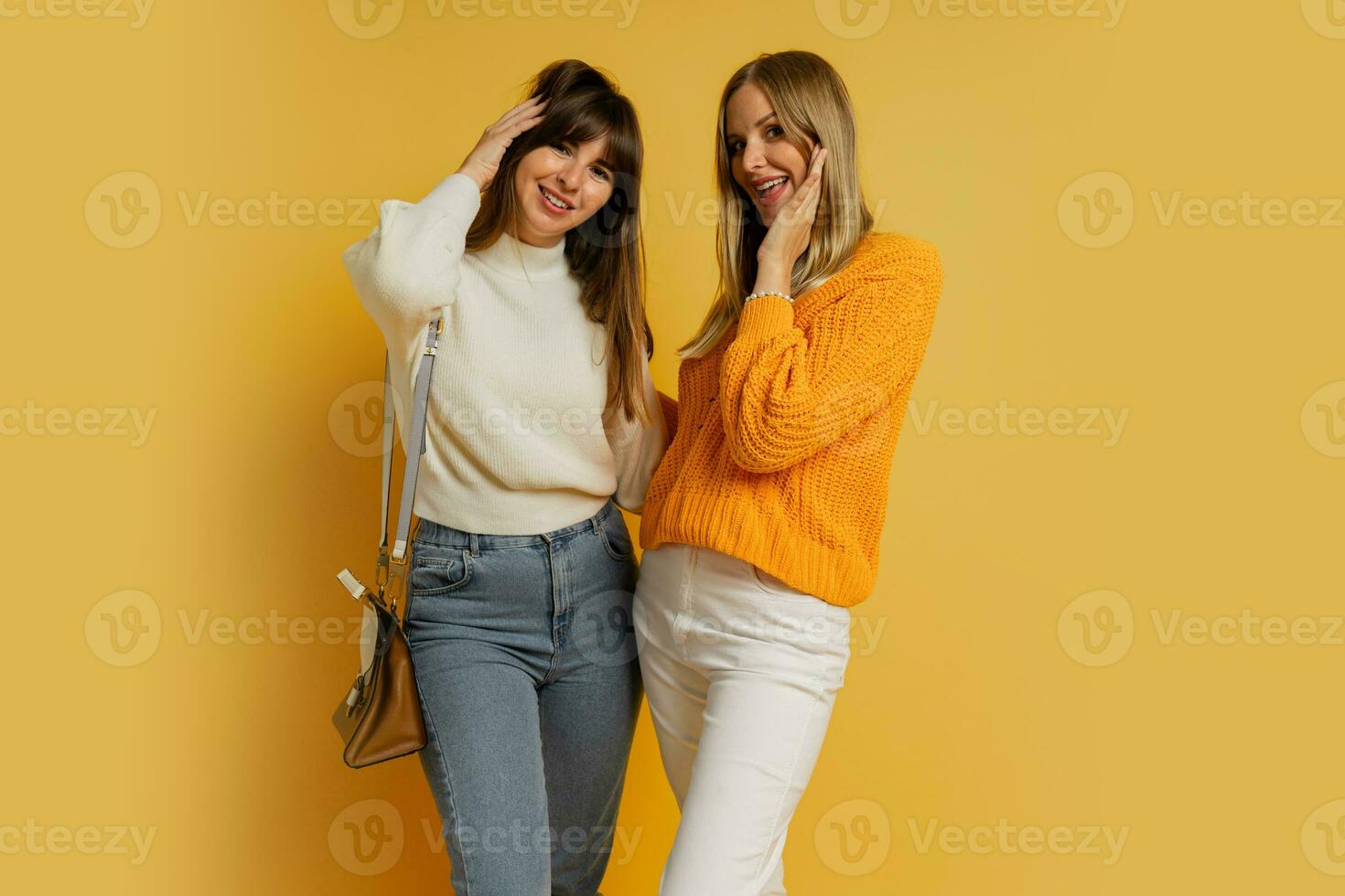 Studio photo of two pretty woman in cozy sweaters posing over yellow background.  Autumn and winter fashion trends.