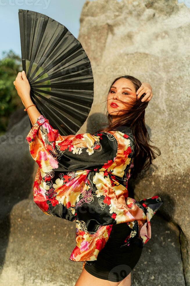 hermosa japonés mujer en elegante kimono con grande ventilador y profesional hacer arriba posando terminado rocas foto