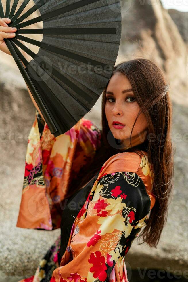Moda foto de elegante asiático mujer en seda kimono participación ventilador y posando terminado rocas en el playa.