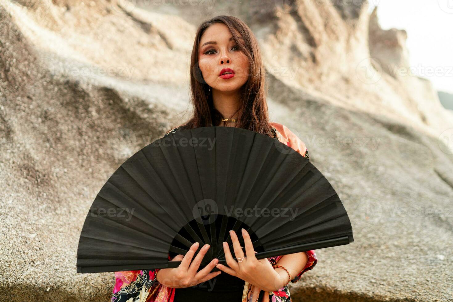 Asian woman  in colorful silk kimono blouse holding black fan and posing on the beach with rocks. photo