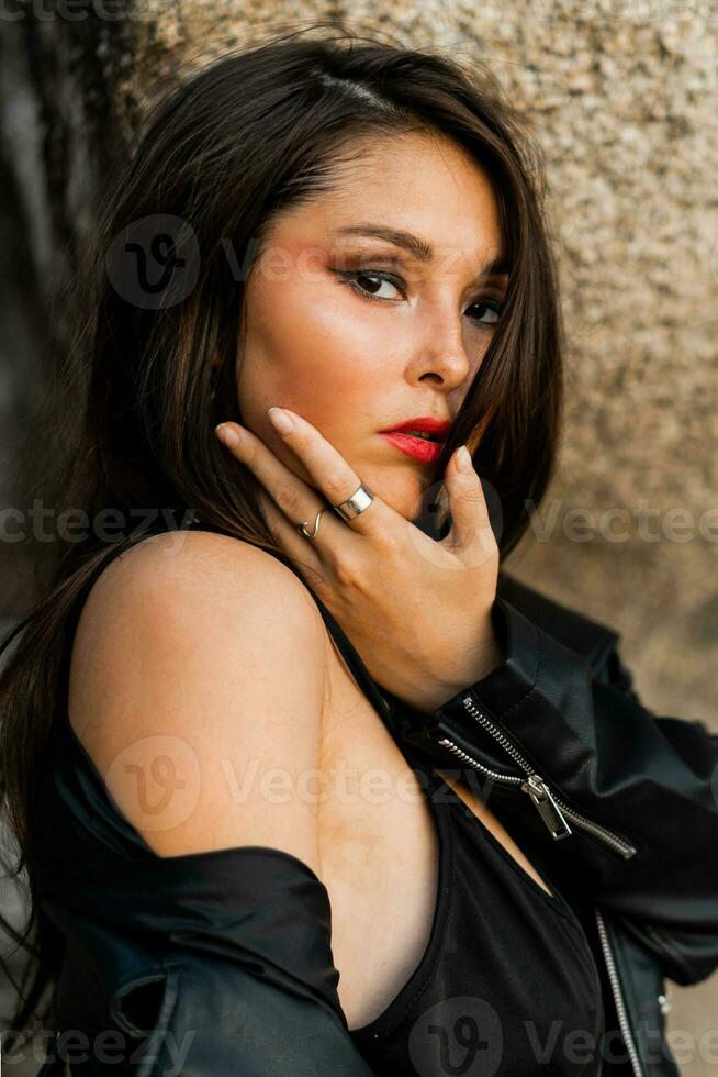 cerca arriba retrato de maravilloso asiático mujer con rojo labios en de moda negro cuero chaqueta y sensual negro vestir posando en rocoso antecedentes. foto