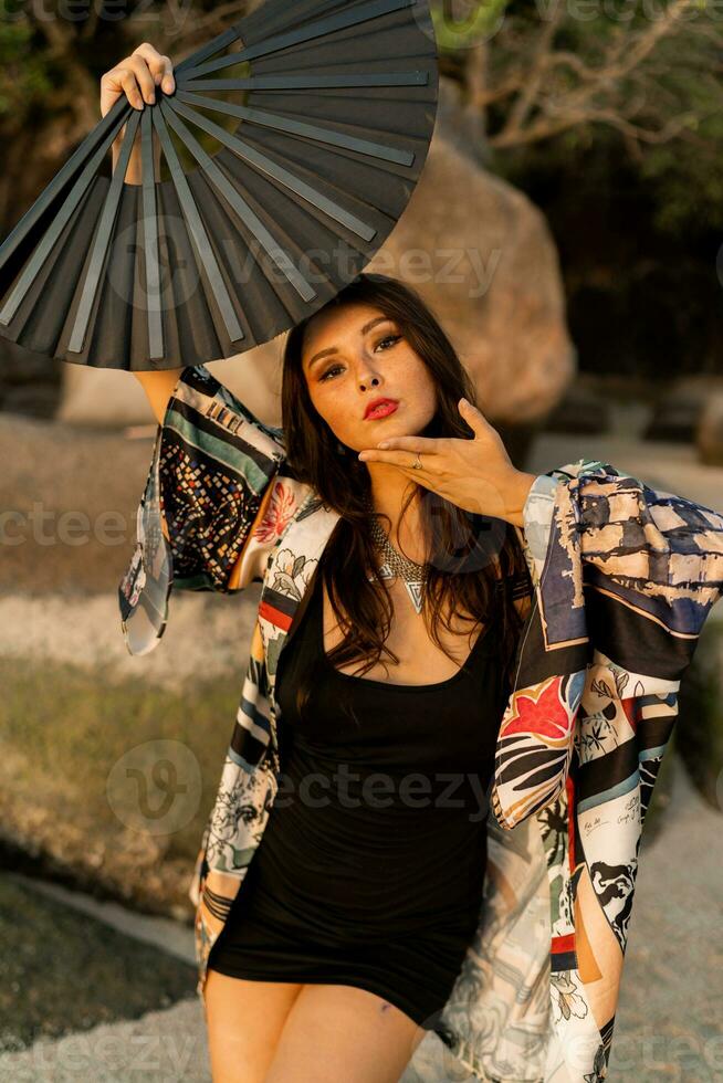 cerca arriba retrato de seductor japonés niña en elegante kimono con grande ventilador y profesional hacer arriba posando en el playa. foto