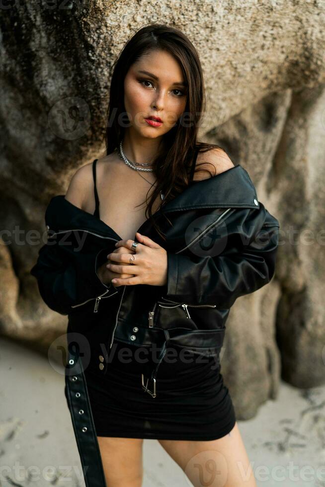 Confident woman  with red lips  in trendy black leather jacket and sensual black dress posing on rocky background. photo