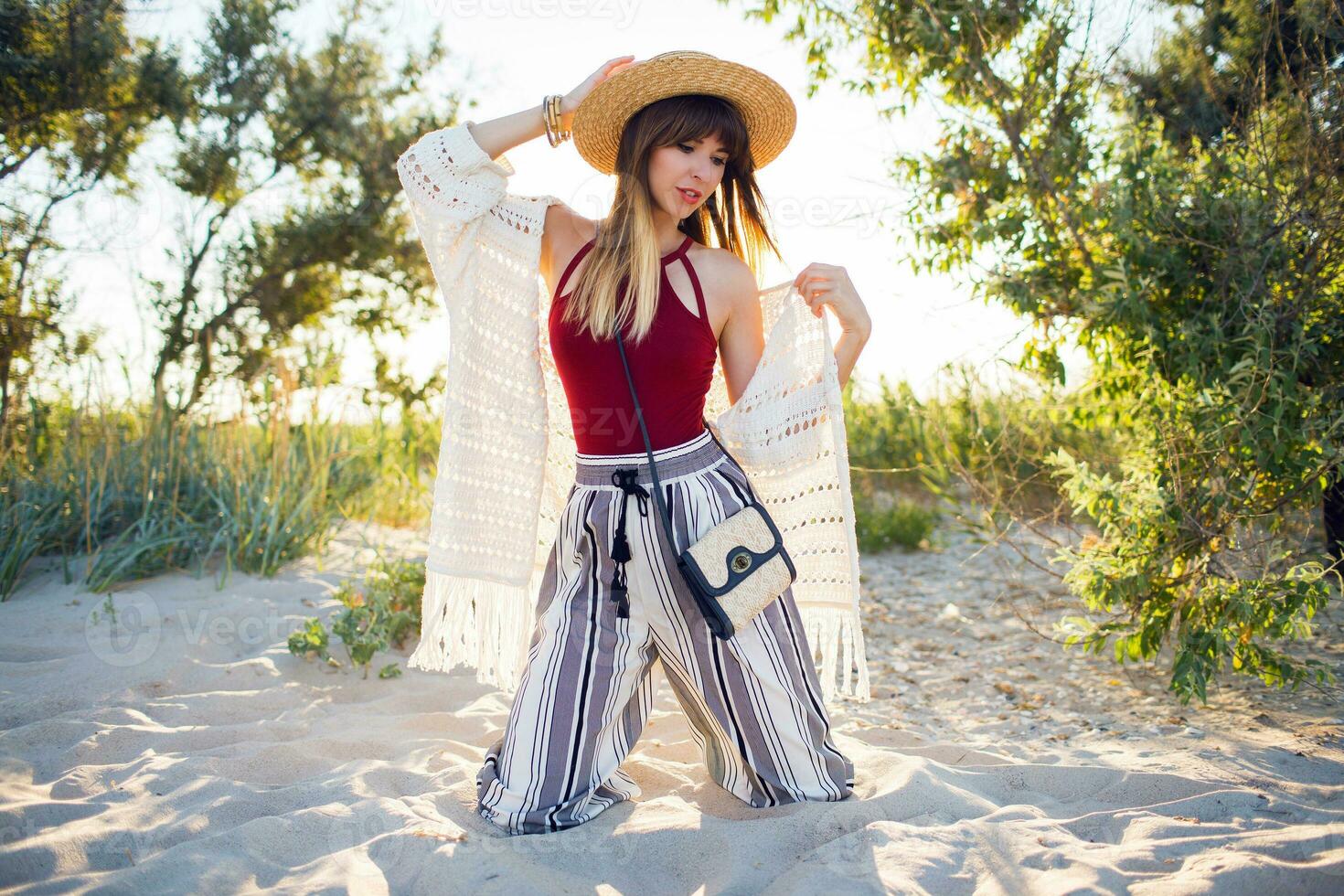 Outdoor summer photo of pretty woman in straw hat posinf on the beach at sunset light.