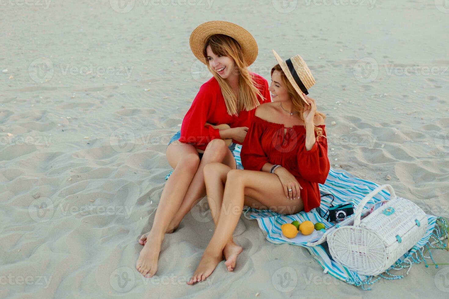 Two pretty women in red summer outfit abd straw hats enjoing picnic on the beach. Summer mood. photo