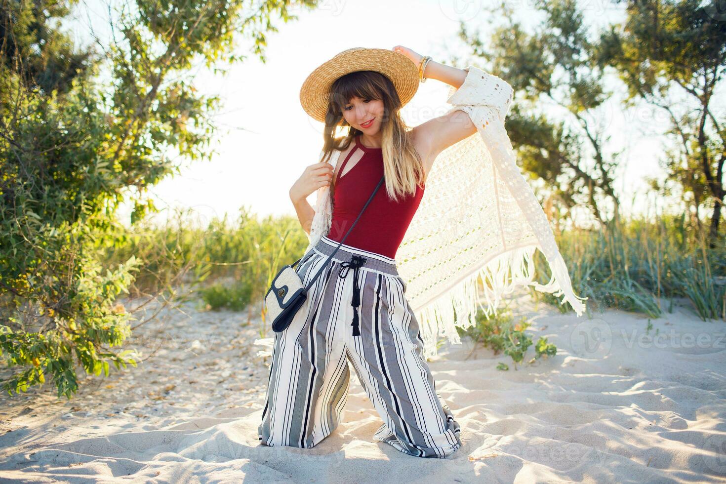 Outdoor summer photo of pretty woman in straw hat posinf on the beach at sunset light.