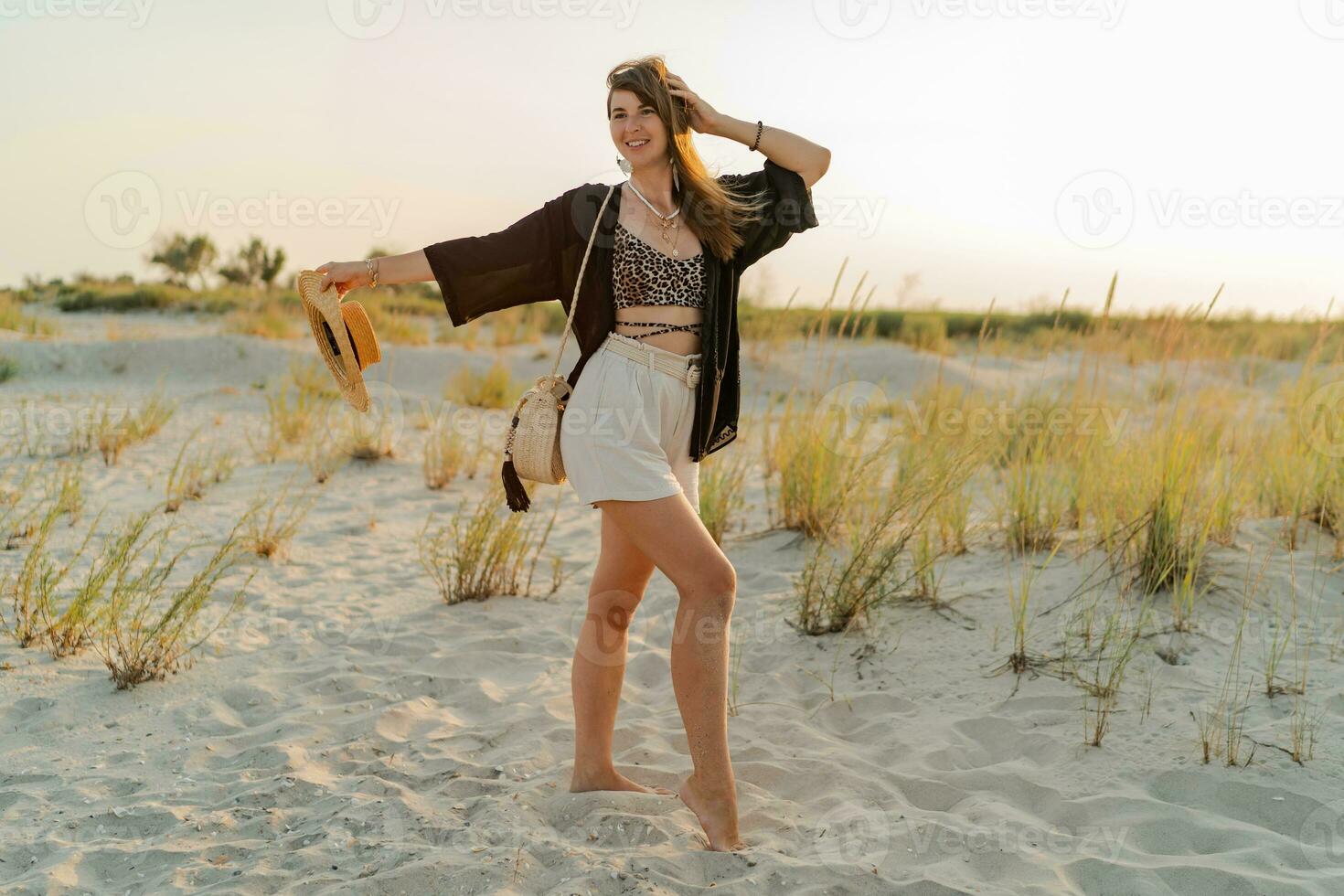 verano foto de contento joven mujer en elegante boho atuendo participación Paja bolso y sombrero posando en el playa. puesta de sol. lleno largo.