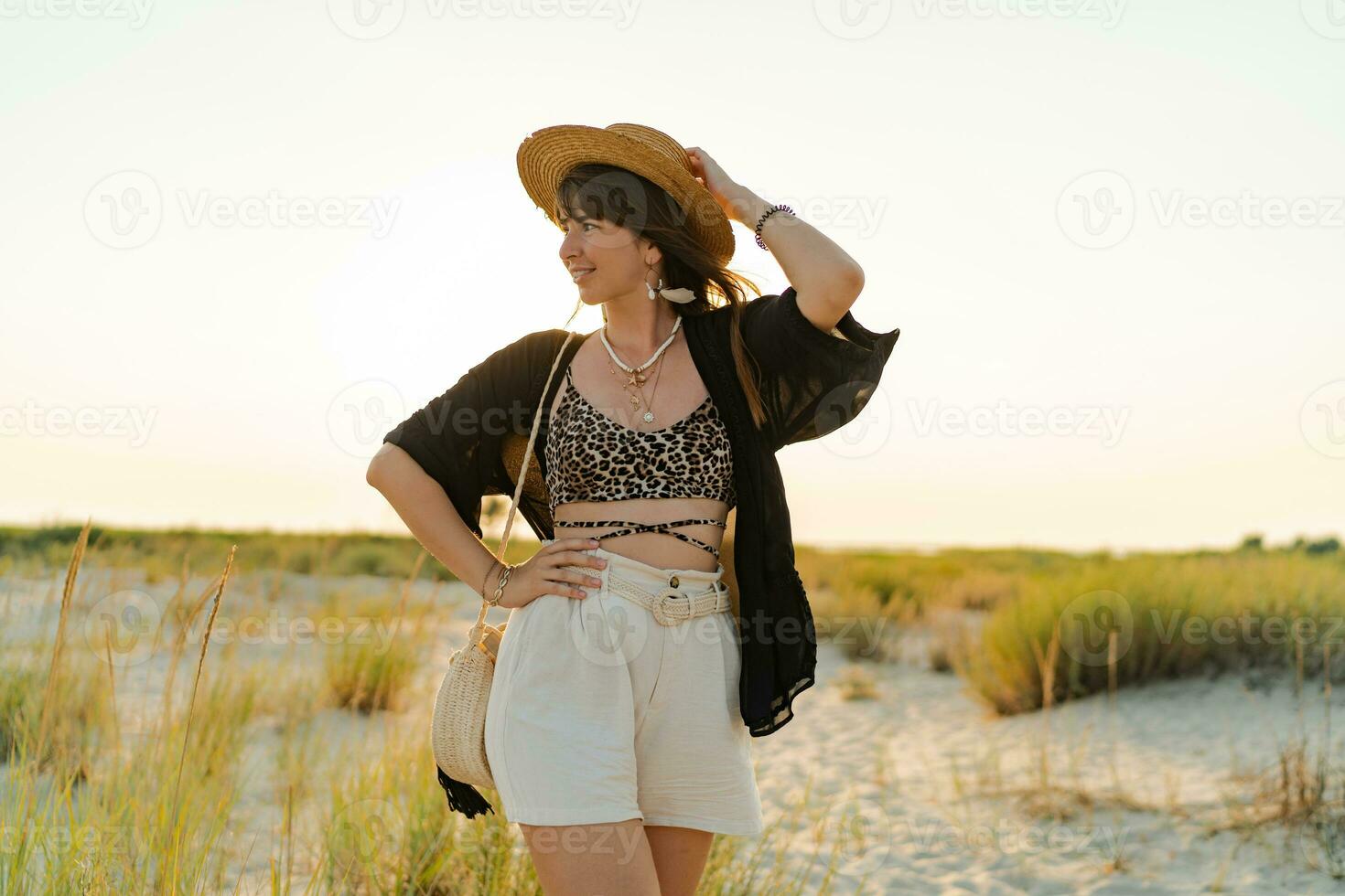 verano foto de contento joven mujer en elegante boho atuendo participación Paja bolso y sombrero posando en el l playa. lleno largo.