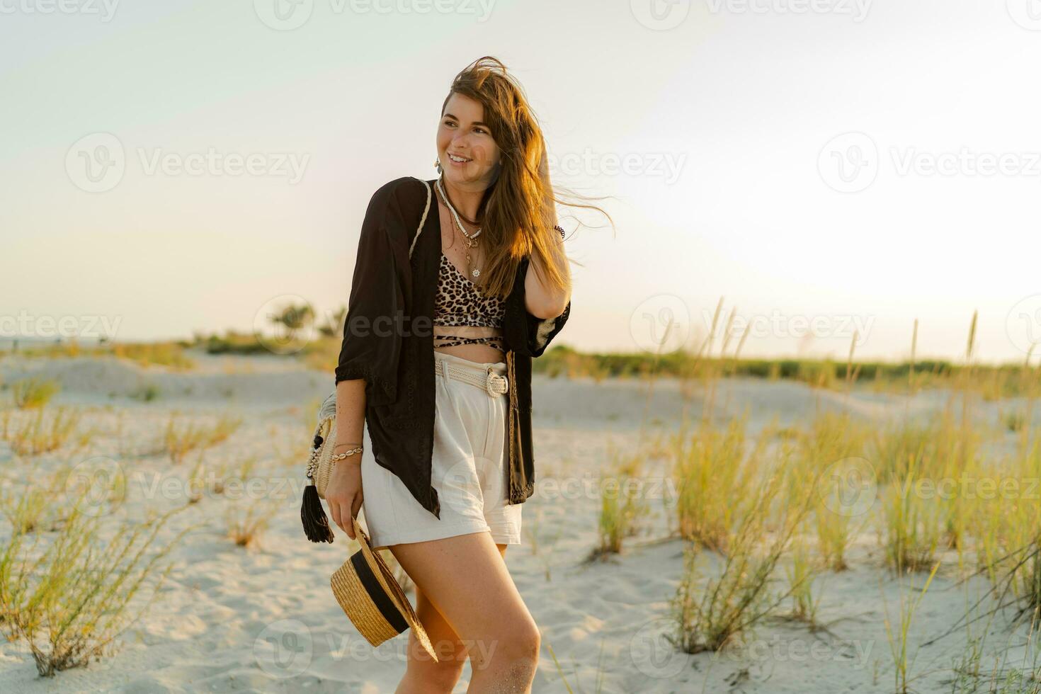 Summer photo of happy young woman in stylish boho  outfit  holding straw bag and hat  posing on  the beach.  Sunset.